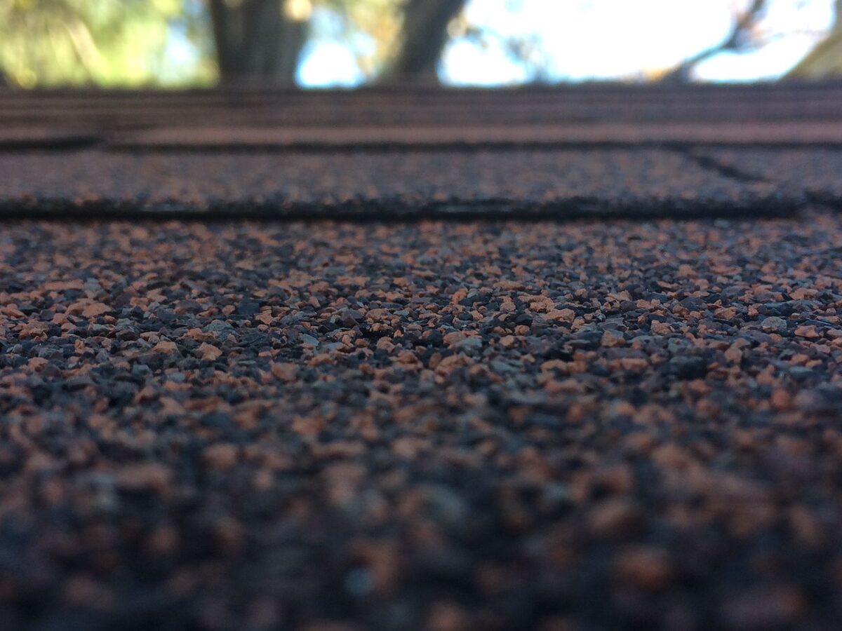 Close-up of weathered asphalt shingles on a residential roof with trees blurred in the background