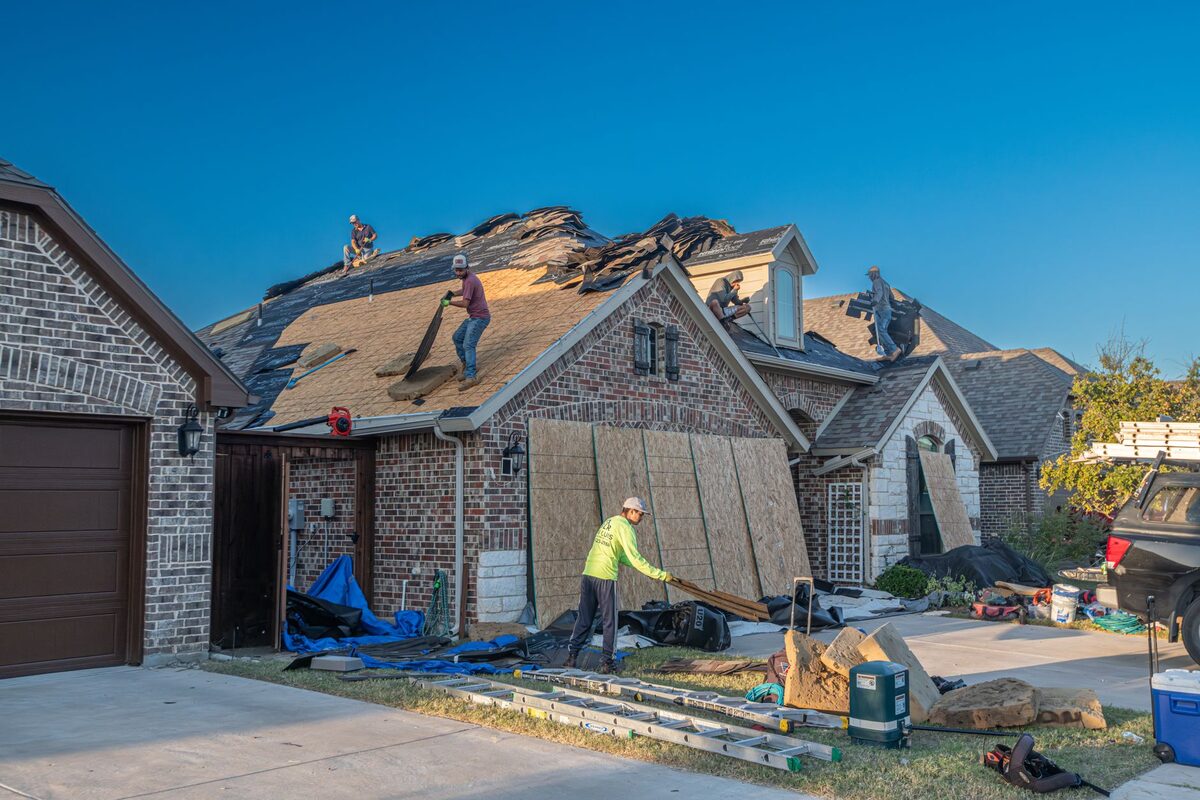 Roofers tearing off old asphalt shingles during a full roof replacement on a brick suburban home
