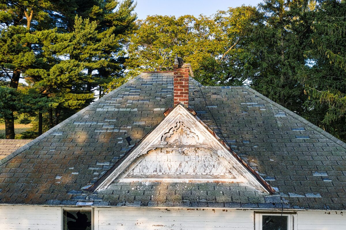 Weathered asphalt shingle roof with patchy wear, streaks and missing pieces around a brick chimney